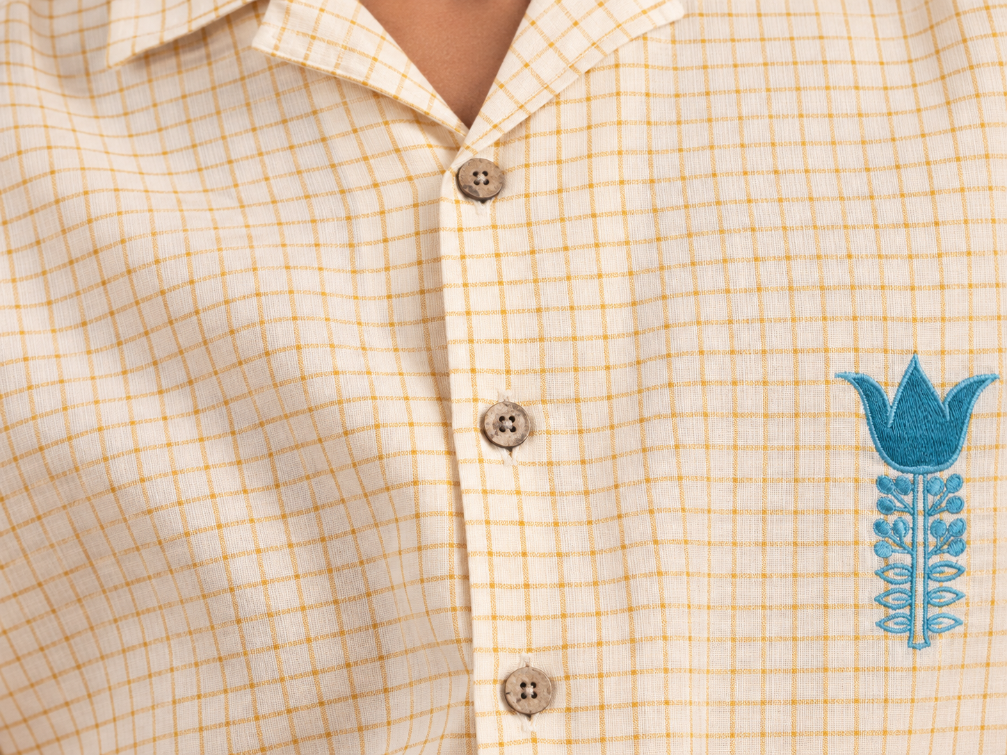 Cream and golden checked shirt with turquoise floral embroidery, button front and short embroidered sleeves Close-up of the open collar and button front on the cream and golden checked shirt Turquoise floral embroidery beside the button front on the checked shirt Short sleeve finished with turquoise leaf embroidery along the border[2]